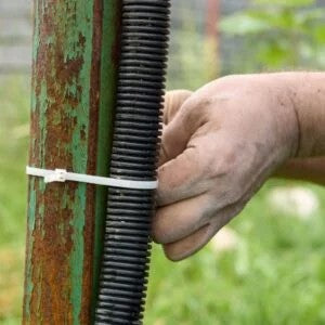 A worker securing a black cable to a rusted pole with a 6-inch black heavy-duty zip tie, from a 100-pack set, ideal for outdoor use with UV protection.